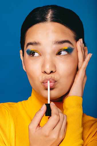 Vibrant young woman applies makeup on her face in a studio, she is wearing graphic eyeliner and lip gloss. Feeling empowered by her makeup look, this woman radiates confidence and self-love as she pouts her lips.