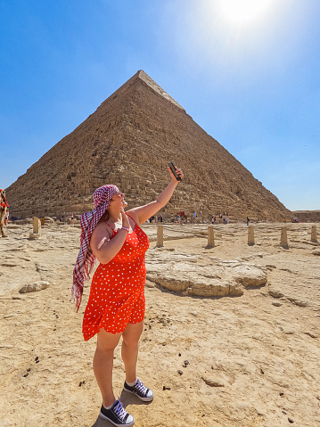 Beautiful young woman having a video call in front of the Pyramids of Giza (Egyptian pyramids) in Cairo, Egypt.
