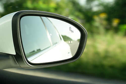 Rear-view mirror, close-up, with the reflection of a white car when the car is moving against the background of green trees. Soft focus. An essential element for driving safety.