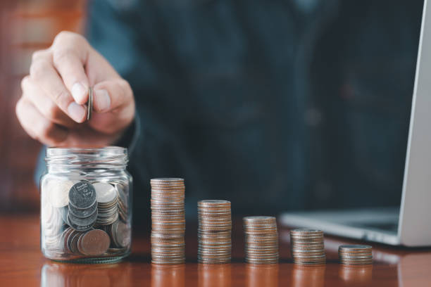 Businessman putting a coin on a glass piggy bank with stack coins, laptop. Saving money, bank deposits, interest collection and taxation concept. Upward trend in business growth, financial management. hombre de negocios poniendo una moneda en una alcancía de vidrio con monedas de pila, computadora portátil. ahorro de dinero, depósitos bancarios, cobro de intereses y concepto de tributación. tendencia al alza en el crecimiento del negocio, gestión f