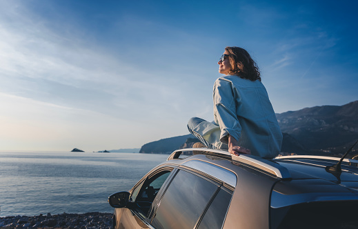 Young happy woman traveler sitting on car roof enjoying sea and sunset view. Summer vacation and adventure concept