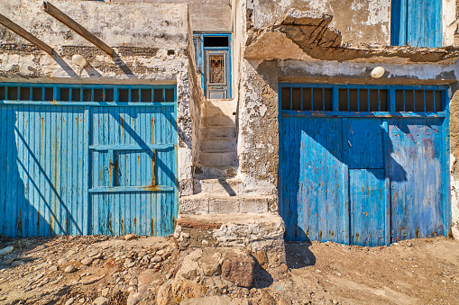 Traditional fisherman house by seafront, Milos island, Greece. Whitewashed and faded walls, painted blue and weathered wooden doors and doorways, boat house or garage, authentic facade, sunny summer day, sunshine and shadow, exterior details.