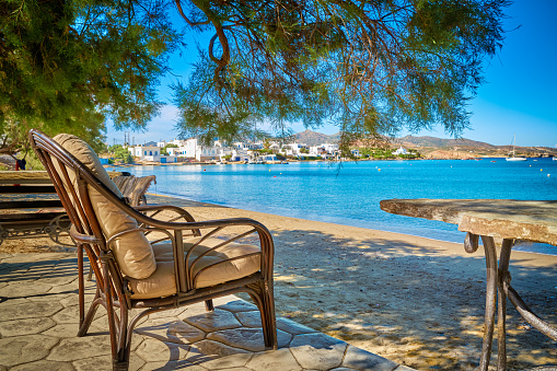 Wicker chair with cushions and stone table in shade of pine tree on beach on sunny summer day and by seafront. Holidays, relax, vacations, distant villages, remote locations, Milos island, Greece.