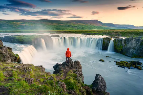 Photo of Godafoss waterfall flowing with colorful sunset sky and male tourist standing on cliff in summer at Iceland