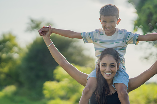 A sweet little boy rides on his Mothers shoulders as they enjoy spending time together outside in the warm summer sun.  They are booth dressed casually and are smiling as they take in the fresh air.