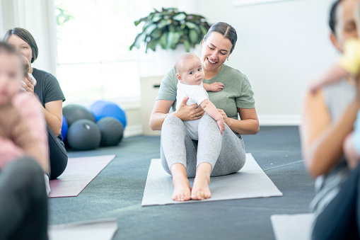 A small group of new parents sit on yoga mats in a studio, with their babies on their laps, as they prepare to participate in a Parent & Baby fitness class.