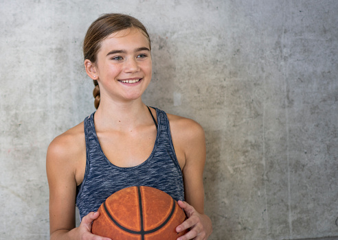 A young female student stands in a gymnasium as she poses for a portrait.  She is wearing athletic wear and holding a basketball.
