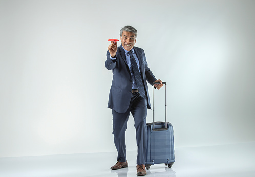 Happy senior businessman with suitcase laughing and flying paper airplane while standing against white background