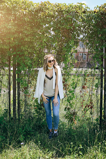 Stylish hipster girl is standing near the ivy leaves wall outdoors. Fashionable photoshoot for young women in white jacket with belt. Blonde short hair and cool glasses. Summer lifestyle photoshoot.