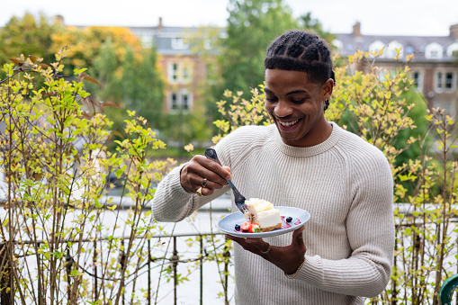Waist-up of a young man standing on the terrace of a restaurant in Newcastle, England. He is eating a slice of cheesecake with strawberries and blackberries with a big smile. He is well dressed.