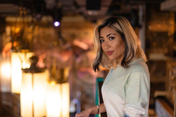 Side view in a charming bar setting, a well-dressed beautiful woman is standing looking at the camera with a confident smile. The backdrop of the bar creates an inviting ambiance. She has a cool attitude with a shaved eyebrow and nose ring.