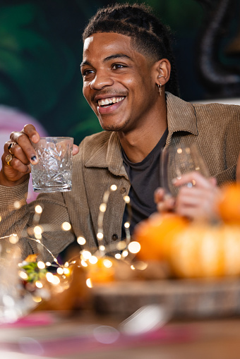 A young man in a charming bar setting, is having a delightful evening. He is gathered around a table with his friends that are out of the shot, his laughter and conversation create an atmosphere of joy and togetherness. He is enjoying a glass of white wine while he waits for his food to arrive, he is having a good time.