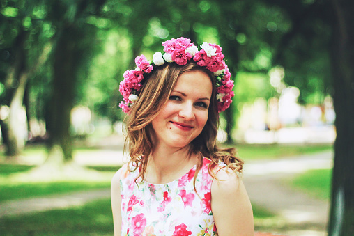 Beautiful girl in pink flower crown smiles you in green summer park. Pink floral dress and emerald trees. Spring woman photo. International women's day and mother's day holidays. Flower chaplet.