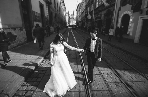 Wedding couple holds hands in old city. Stone walls of ancient town and tram railways. Rustic bride with hair down and groom in grey suit and bow tie. Romantic love in vintage street. Black and white.