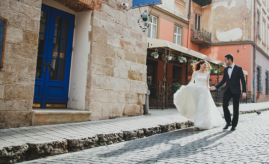 Wedding couple runs in the old city. Blue doors and cafe in ancient sunny town on background. Rustic bride with hair down and groom in grey suit and bow tie. Romantic love in vintage atmosphere.