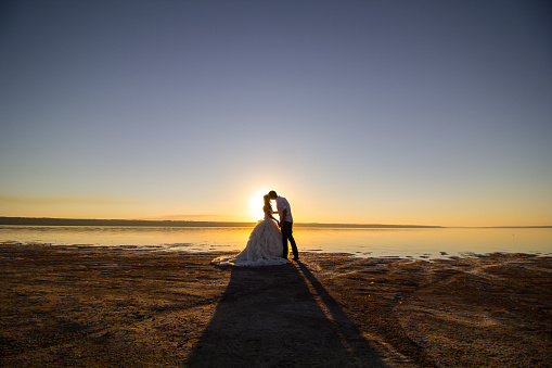 Wedding couple kissing on the sea beach watching sunset. Sunny summer photo. Bride with hair down in off shoulder dress with train. Ocean romantic ceremony. Seaside sunrise love story. Honeymoon.