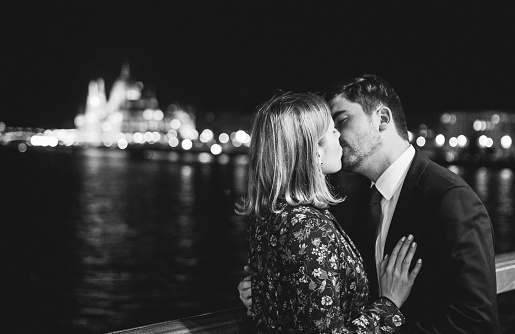 Couple in love is kissing and hugging with a view on Budapest parliament. Night city lights in european city. Man and woman silhouettes near river panoramic view. Black and white stylish fashion photo