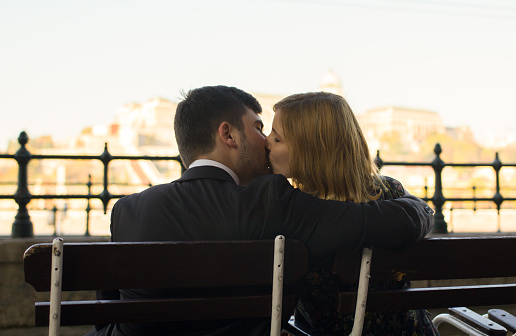 Couple in love is kissing and hugging on the wooden bench in the morning city. man and woman in black clothes are having great time.