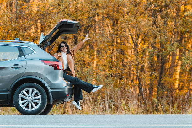 una donna sorridente siede nel bagagliaio di un'auto. sosta per il viaggio su strada - car insurance women driving foto e immagini stock