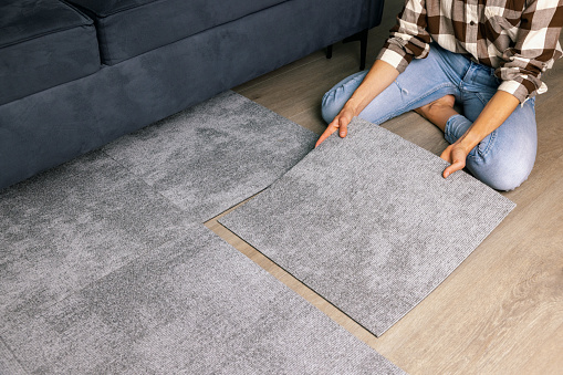 woman laying self adhesive carpet tiles on floor in living room at home