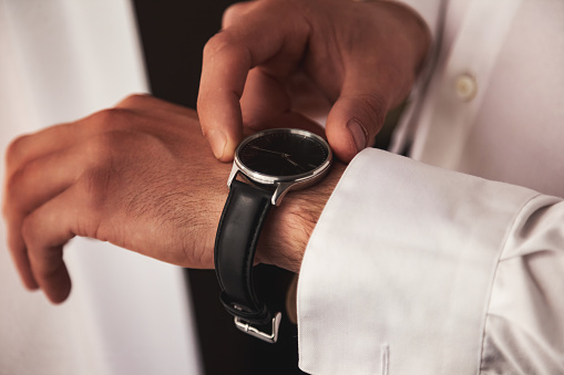 Close up of hands confident man businessman in white shirt putting on watch at window in living room. Close-up guy puts wristwatch on his hand. Concept of business meeting. Copy ad text space poster
