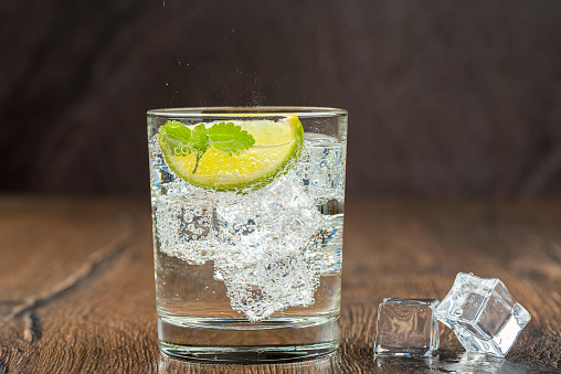 Mineral refreshing water in a glass with ice, mint leaves and lime slice. Dark wooden surface. Artificial ice cubes.