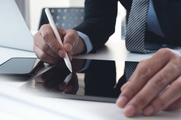 istock Businessman in black suit using stylus pen signing e-document on digital tablet via mobile app and working on laptop computer on table at office. Business man suring the internet, close up 1629669786