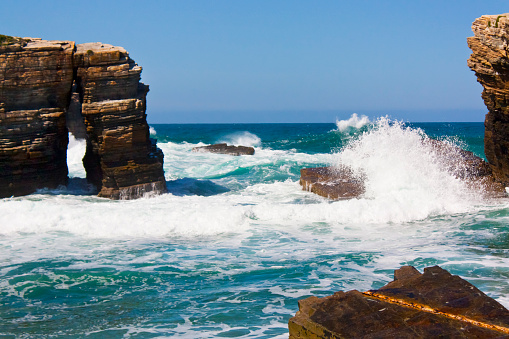 Wild    beach, breaking waves , rocky coastline, natural arch, horizon over water, clear sky. Cliffs and bay near Cathedrals beach, Ribadeo , A Mariña, Lugo province, Galicia, Spain.