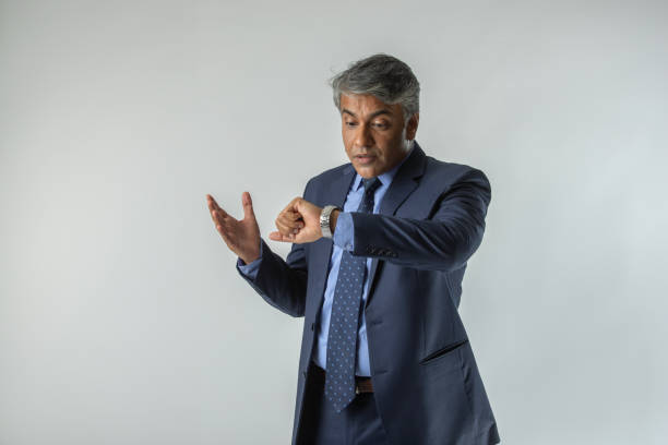 Worried senior businessman dressed in elegant suit checking time on wristwatch while standing against white background