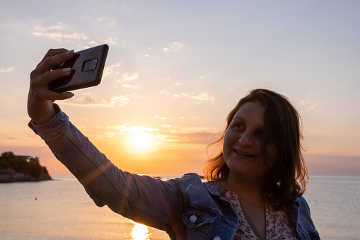 A young woman with a phone in her hands takes a selfie against the background of the rising sun.