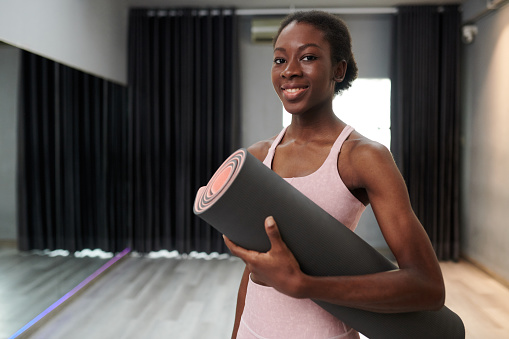 Smiling young Black woman standing in her yoga studio with mat in hands