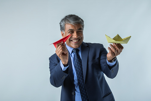 Smiling Indian businessman holding paper boat and paper airplane while standing against white background