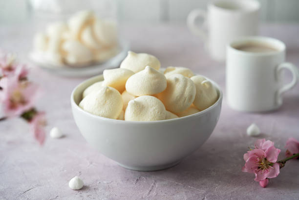 istock Small meringues in a white bowl over light pink background decorated with spring peach tree flowers, two cup of coffee behind 1629738177
