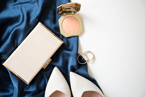 The beige women’s small handbag, pink powder and beige women's shoes are on a cocktail blue dress on a white background. Copy space.