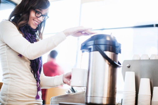 A beautiful young adult hipster woman smiles as she fills a mug of hot drip coffee from a stainless steel carafe, bright white light coming in through the cafe windows in the background.  Horizontal with copy space.