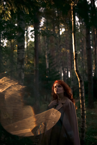 Beautiful dreamy red-haired woman in beige coat in autumn forest sunset time. Sunset light on face, backlight from a flashlight. Lens flare effect.