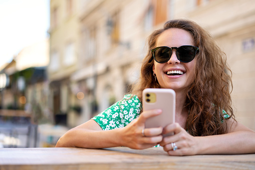 Curly young adult woman wearing sunglasses sits in side walk cafe using her mobile phone and laughing.