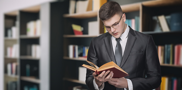 A male lawyer or manager reads a textbook on the background of a shelf with literature.
