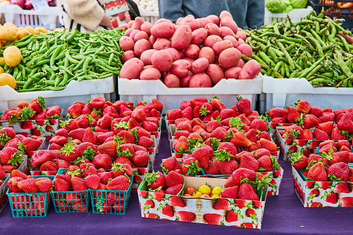 Image of Strawberries in containers on table with vegetables in crates behind them and line of people