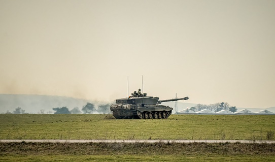 Salisbury, United Kingdom – March 22, 2023: An olive-green military tank driving across an expansive green field, with a large brick building in the background