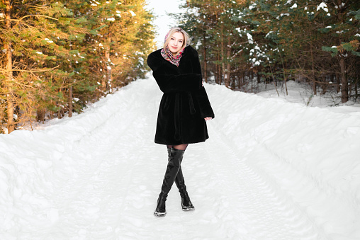 Woman in a black fur coat in a headscarf in leather boots walks along the road in a winter coniferous forest.