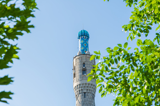 Close-up view of  stone minaret with blue roof of old Saint Petersburg Mosque. Natural frame from green leaf branches. Clear blue sky. Religious architecture. Muslim culture theme.