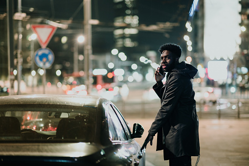 An urban African American man is entering a taxi car at night while talking on the phone. A man is entering car while having a phone call.
