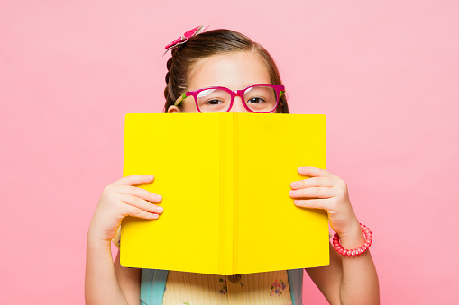 Cute little girl wearing glasses hiding behind a book while reading and studying at school against a pink studio background