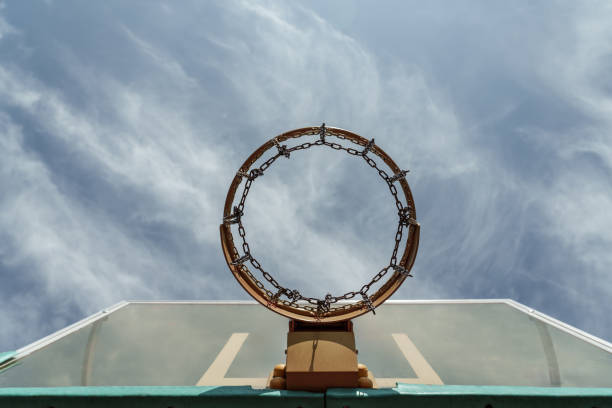A photo of a basketball hoop with an orange rim and a metal chain net. The photo is taken from below the hoop and shows the blue sky and the white clouds through the net.