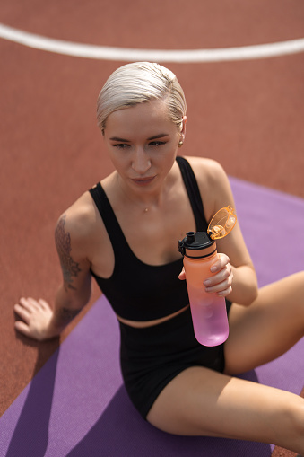 A blonde woman with a short haircut is sitting on a purple yoga mat on a red running track holding a pink water bottle and wearing a black sports bra and shorts. She has a tattoo on her right arm