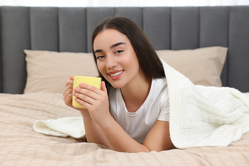 Happy young woman under plaid holding yellow ceramic mug on bed