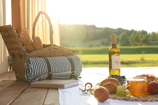 Romantic date. Wicker basket, glass of wine and snacks for picnic on white blanket outdoors