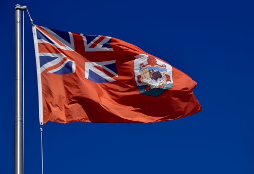 Hamilton, Bermuda: Bermudian flag against blue sky - The flag of the British Overseas Territory of Bermuda as a red ensign was first adopted in 1910. It is a British Red Ensign with the Union Flag (Union Jack) in the upper left corner, and the coat of arms of Bermuda in the lower right. The coat of arms of Bermuda displays a red lion holding a shield that has a depiction of a wrecked ship upon it. The red lion is a symbol of Great Britain. The wrecked ship is the Sea Venture, a 17th-century English sailing ship, part of the Third Supply mission to the Jamestown Colony, that was wrecked in Bermuda in 1609.