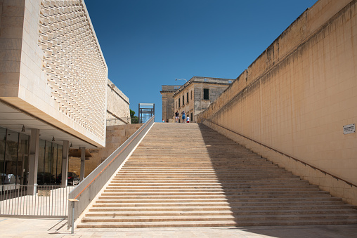 View of the grand stone staircase in the old town of Valetta, Malta. Part of the parliament building on the side. The sky is blue.